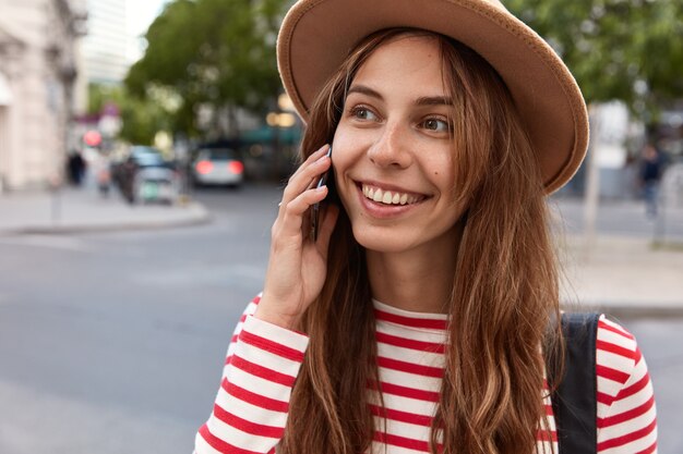 Vue recadrée de l'heureuse femme du Caucaisan avec un doux sourire, parle via téléphone portable, regarde de côté