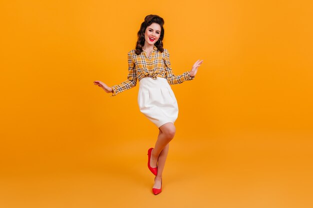 Vue sur toute la longueur de la femme frisée gracieuse en jupe blanche. Pin-up en chemise à carreaux dansant sur fond jaune.