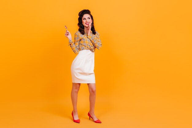 Vue sur toute la longueur d'une fille brune bien habillée avec une sucette. Photo de Studio de belle femme pin-up posant sur fond jaune.