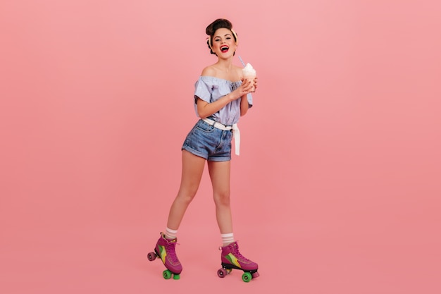 Photo gratuite vue sur toute la longueur d'une fille heureuse avec un milkshake. pinup femme sur patins à roulettes tenant un cocktail sur fond rose.