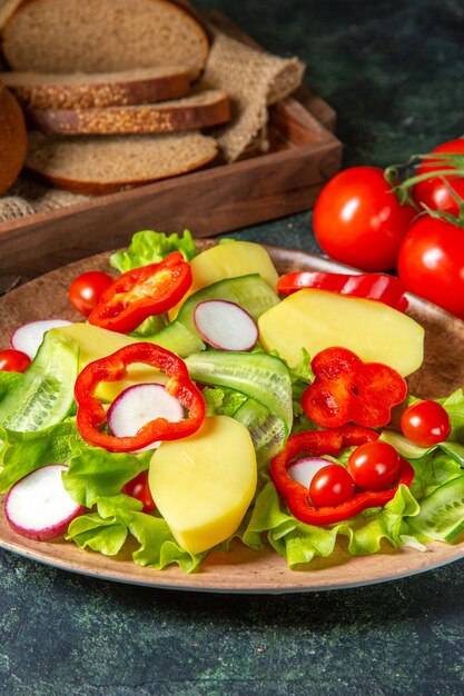 Vue verticale de couper du pain noir frais sur une serviette dans une boîte en bois brun et haché des légumes frais sur une assiette sur la surface des couleurs de mélange sombre