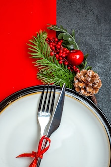 Vue verticale du fond de noël avec des couverts sertie de ruban rouge sur une assiette à dîner accessoires de décoration branches de sapin sur une serviette rouge