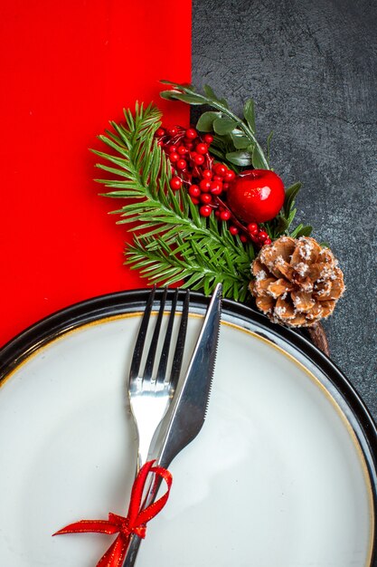 Vue verticale du fond de Noël avec des couverts sertie de ruban rouge sur une assiette à dîner accessoires de décoration branches de sapin sur une serviette rouge