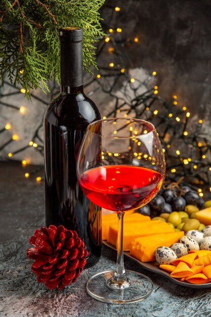 Vue verticale du vin rouge sec dans un verre et dans une bouteille à côté d'un en-cas et d'un cône de conifère sur fond gris