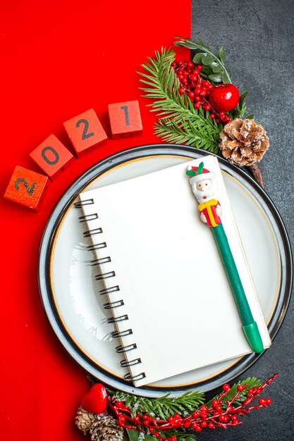 Vue verticale de fond de nouvel an avec cahier à spirale sur assiette à dîner accessoires de décoration branches de sapin et numéros sur une serviette rouge sur un tableau noir