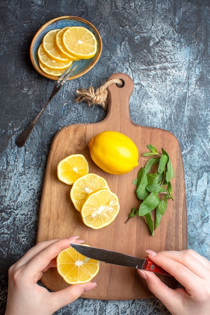 Vue verticale d'une main coupant des citrons frais et de la menthe sur une planche à découper en bois sur fond sombre