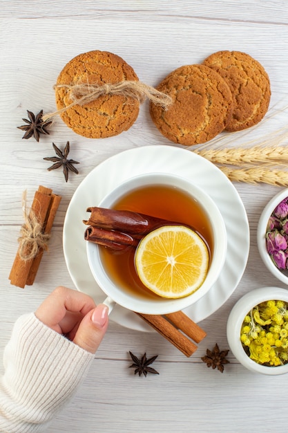 Vue verticale d'une main de femme tenant une tasse de thé noir avec du citron vert à la cannelle et divers biscuits à base de plantes sur fond blanc