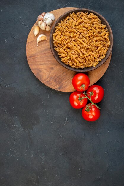 Vue verticale de pâtes crues dans un bol brun ails sur des pointes de planche de bois et des tomates avec des tiges sur fond noir
