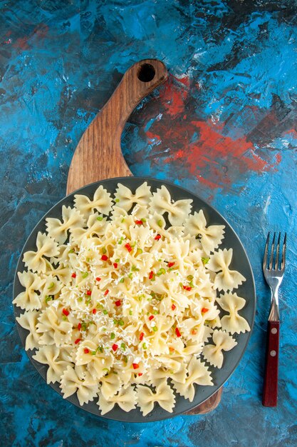 Vue verticale des pâtes farfalle italiennes crues avec des légumes sur une plaque noire sur une planche à découper en bois et une fourchette sur fond bleu