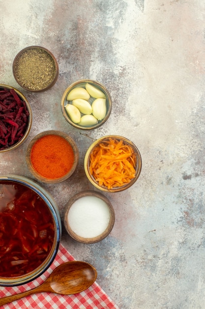 Vue verticale d'un pot bleu avec une délicieuse soupe de bortsch et différentes épices et légumes cuillère en bois sur une serviette dénudée rouge sur fond coloré