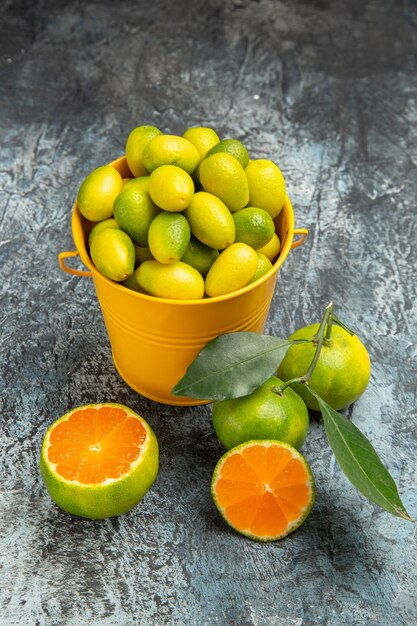 Vue verticale d'un seau jaune plein de mandarines vertes fraîches et coupées en deux mandarines sur fond gris