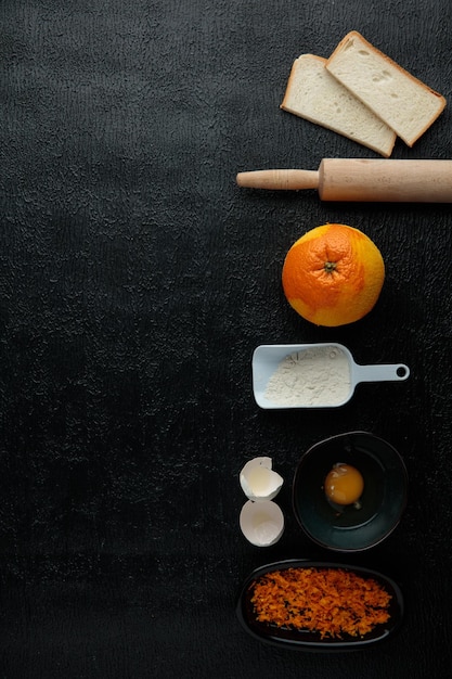 Vue verticale des tranches de pain orange jaune d'oeuf zeste d'orange cuillère à mesurer avec farine avec rouleau à pâtisserie et coquille d'oeuf sur fond noir