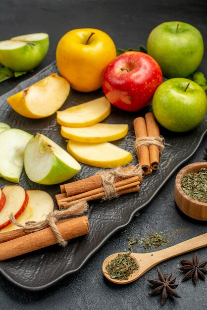 Vue verticale de tranches vertes jaunes et rouges et de pommes fraîches entières sur un plateau noir et de citrons verts à la cannelle sur une table sombre