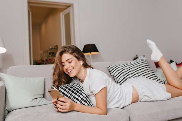 Winsome femme allongée sur un canapé avec des coussins et appréciant la chanson