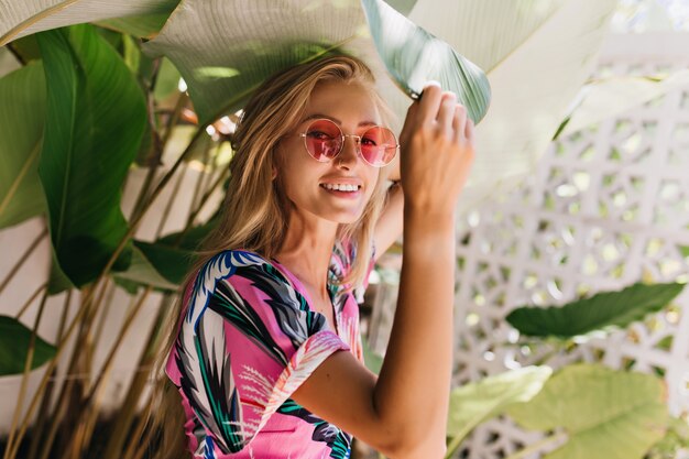 Winsome femme aux cheveux longs dans des lunettes de soleil roses posant à côté des plantes.