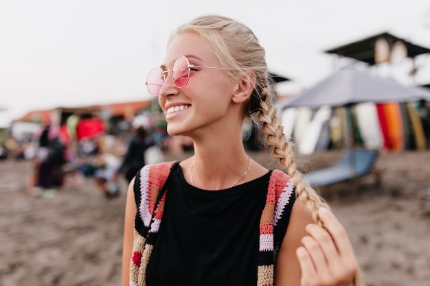 Winsome femme bronzée posant avec le sourire à la plage. Portrait en plein air de femme blonde à lunettes roses jouant avec ses tresses.