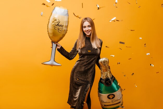 Winsome jeune femme au visage heureux expressiin en attente de fête d'anniversaire. Portrait intérieur d'une femme blonde galbée avec une bouteille de champagne et un verre à vin.