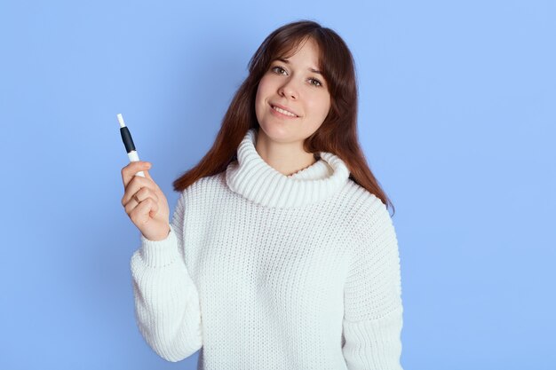 Winsome vaping jeune femme sur bleu, dame de bonne humeur regarde la caméra et tient e ciggy dans sa main, vêtue d'une tenue décontractée, a les cheveux noirs.