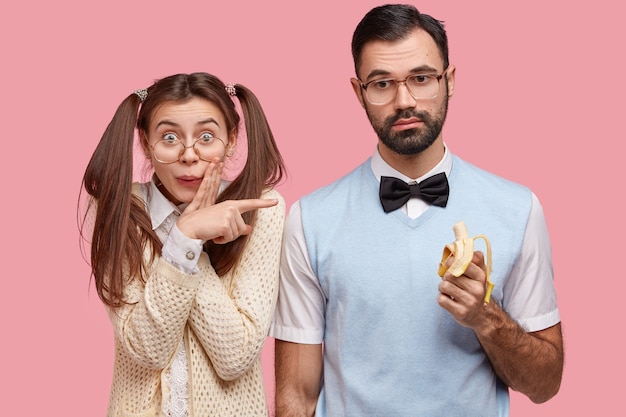 Wonk femelle drôle positif a les cheveux noirs peignés en deux queues de cheval, pointe au meilleur ami qui mange de la banane, vêtu de vêtements élégants