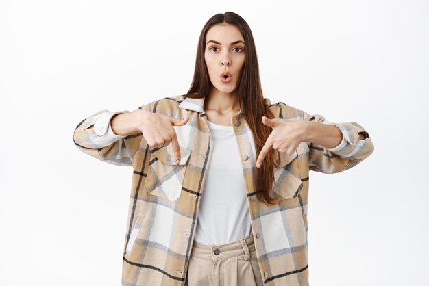 Wow cool offre promotionnelle ci-dessous Une jeune femme intriguée pointe vers le bas et regarde avec un visage surpris et intéressé en vérifiant la nouvelle offre promotionnelle de la boutique debout sur fond blanc