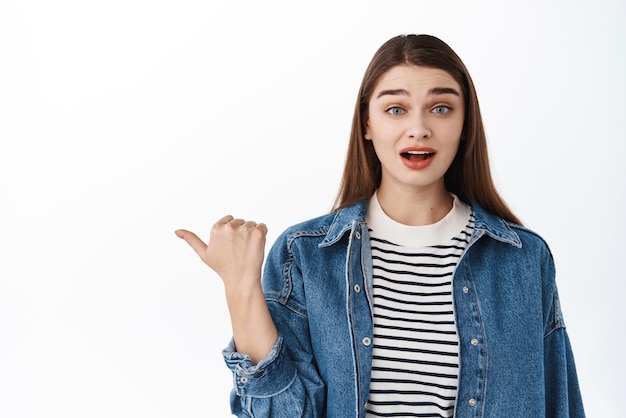 Wow est-ce vrai Intrigué jolie fille haletant regardant intéressé pointant le pouce à gauche sur l'espace de copie posant une question sur le produit de spectacle de logo de bannière intéressant debout sur fond blanc