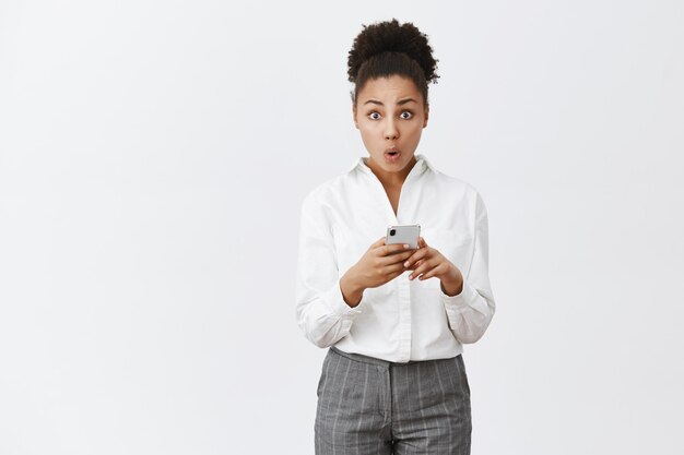 Wow nouvelles fonctionnalités après la mise à jour. Portrait de femme afro-américaine mignonne curieuse en chemise blanche élégante et pantalon, tenant smarpthone, regardant impressionné par les lèvres pliées et le regard surpris