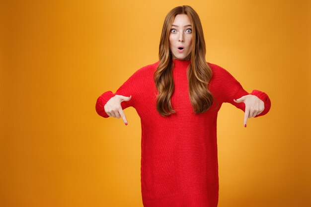 Wow quelle promotion géniale. Portrait d'une femme rousse mignonne impressionnée et surprise en robe tricotée chaude et élégante pliant les lèvres avec étonnement pointant vers le bas et regardant la caméra