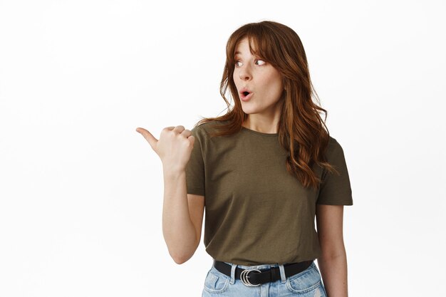 Wow, regarde ça. Jeune femme impressionnée pointant vers la gauche et regardant la publicité debout sur blanc