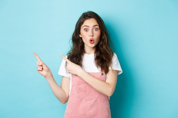 Wow, regarde ça. Le modèle féminin brune étonnée plisse les lèvres et a l'air intrigué, pointant du doigt quelque chose d'intéressant, montrant une bannière, fond bleu