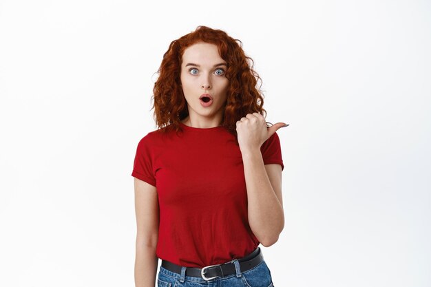 Wow regarde là. Fille rousse excitée et surprise pointant le pouce vers la bannière de droite, haletant étonné, montrant l'offre promotionnelle de côté sur blanc