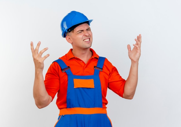 Avec les yeux fermés confus jeune constructeur masculin portant l'uniforme et un casque de sécurité répand les mains sur le blanc