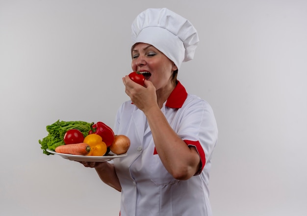 Avec les yeux fermés femme d'âge moyen cuisinier en uniforme de chef tenant des légumes sur la plaque mord la tomate avec copie espace