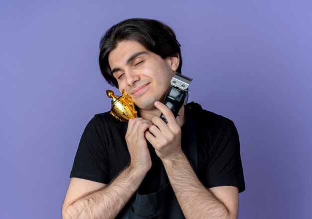 Avec les yeux fermés heureux jeune beau coiffeur masculin en uniforme tenant une tondeuse à cheveux et coupe gagnant autour de la gorge isolé sur bleu