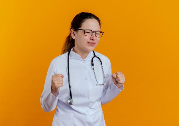 Avec les yeux fermés heureux jeune femme médecin portant une robe médicale et un stéthoscope avec des lunettes montrant oui geste isolé