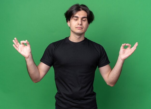 Avec les yeux fermés jeune beau mec portant un t-shirt noir montrant le geste de méditation isolé sur le mur vert