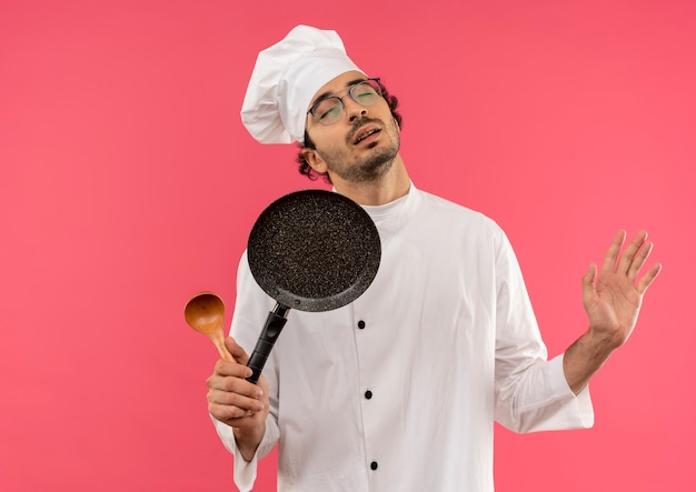 Avec les yeux fermés jeune cuisinier homme portant un uniforme de chef et des verres tenant une poêle avec une cuillère et une cuillère à main sur rose