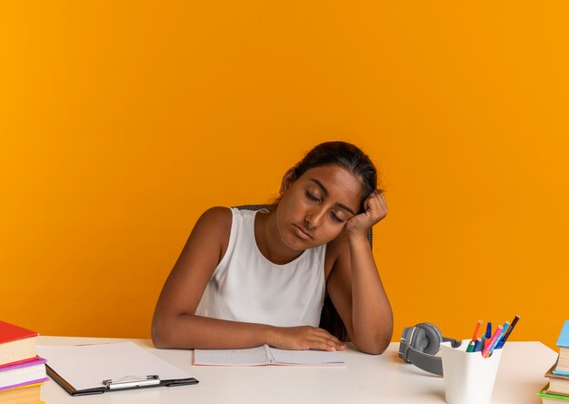 Avec les yeux fermés jeune écolière fatiguée assis au bureau avec des outils scolaires mettant la tête en main sur l'orange