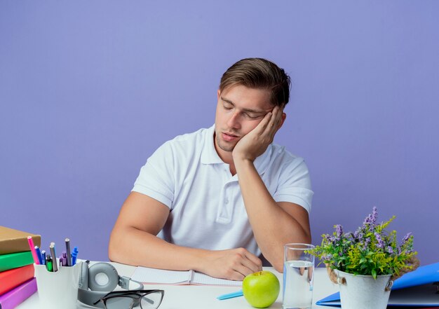 Avec les yeux fermés jeune étudiant masculin beau assis au bureau avec des outils scolaires montrant le geste de sommeil isolé sur bleu