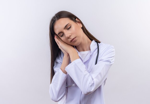 Avec les yeux fermés jeune fille portant une robe médicale stéthoscope montrant le geste de sommeil sur blanc isolé