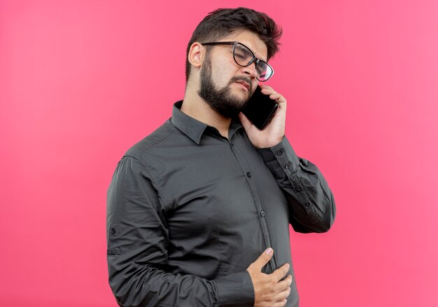 Avec les yeux fermés, jeune homme d'affaires portant des lunettes parle au téléphone isolé sur rose