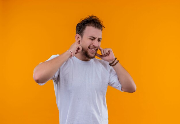 Avec les yeux fermés jeune homme portant un t-shirt blanc oreilles fermées sur mur orange isolé