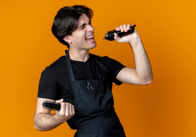 Avec les yeux fermés joyeux jeune homme beau coiffeur en uniforme tenant une tondeuse à cheveux avec peigne et chant isolé sur orange