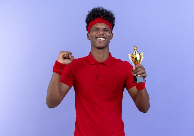 Photo gratuite avec les yeux fermés joyeux jeune homme sportif afro-américain portant bandeau et bracelet tenant la coupe du gagnant et montrant oui geste isolé sur bleu