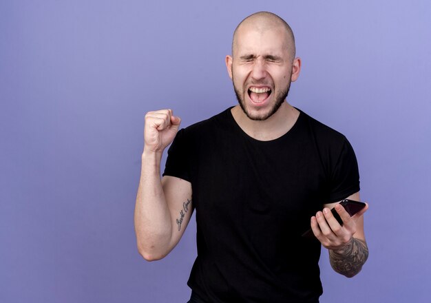 Avec les yeux fermés joyeux jeune homme sportif tenant le téléphone et montrant oui geste isolé sur violet