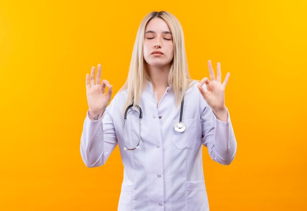 Avec les yeux fermés médecin jeune fille portant un stéthoscope en robe médicale montrant le geste okey avec les deux mains sur un mur jaune isolé