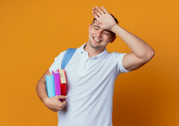 Avec les yeux fermés souriant jeune beau étudiant masculin portant un sac à dos tenant des livres