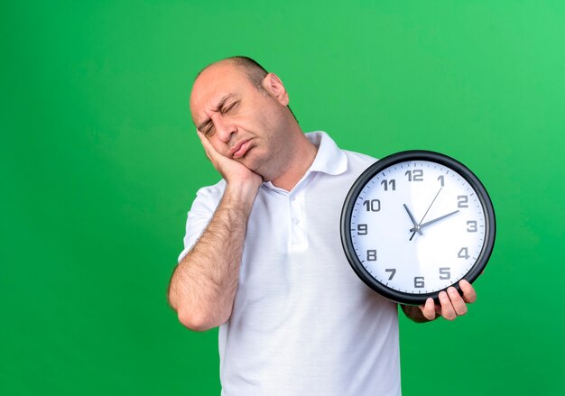 Avec les yeux fermés triste homme mûr occasionnel tenant horloge murale et mettant la main sur la joue isolé sur mur vert