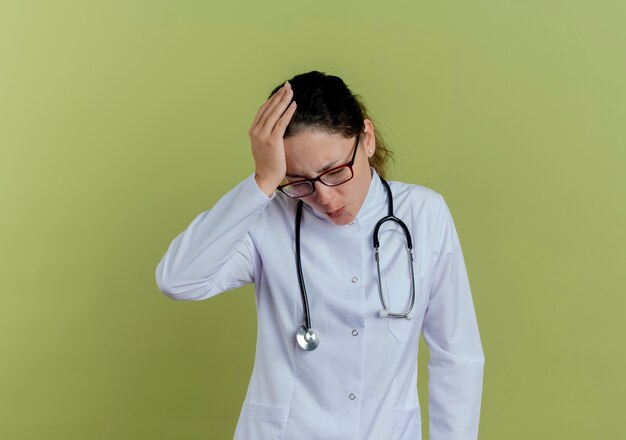 Avec les yeux fermés triste jeune femme médecin portant une robe médicale et un stéthoscope avec des lunettes mettant la main sur la tête isolée sur un mur vert olive