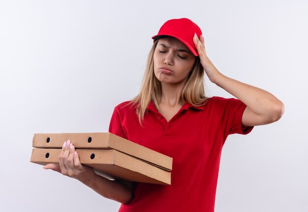 Avec les yeux fermés triste jeune livreuse portant un uniforme rouge et une casquette tenant des boîtes à pizza et mettant la main sur la tête isolé sur blanc