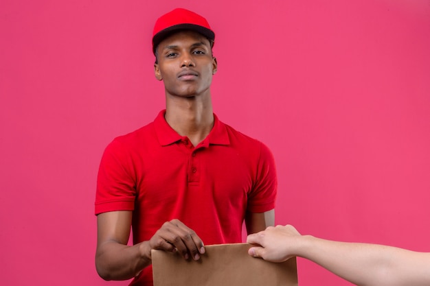 Young african american delivery man wearing red polo shirt and cap à confiant donnant boîte de papier au client sur rose isolé